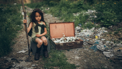 A homeless girl is sitting on a garbage dump next to a suitcase with flowers inside.