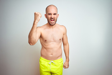 Young shirtless man on vacation wearing yellow swimwear over isolated background angry and mad raising fist frustrated and furious while shouting with anger. Rage and aggressive concept.