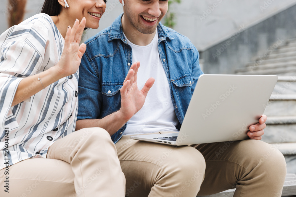 Wall mural Happy young amazing loving couple business people colleagues outdoors outside on steps using laptop computer waving talking with family or friends.