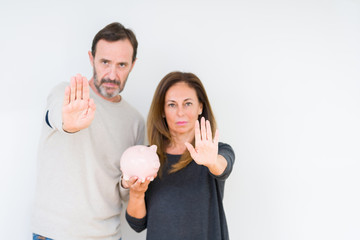 Middle age couple holding piggy bank over isolated background with open hand doing stop sign with serious and confident expression, defense gesture