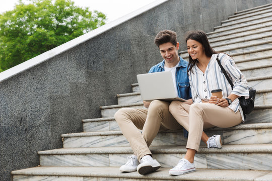 Image of optimistic couple with paper cup smiling and looking at laptop while sitting on stairs outdoors