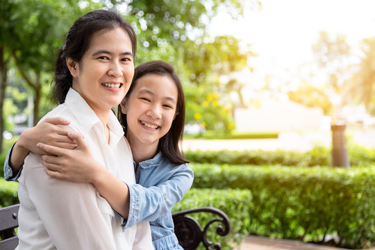 Happy beautiful asian adult woman and cute child girl sitting on bench with hugging ,smiling in garden,love of mother with her little daughter in outdoor park ,mother’s day, family, love concept