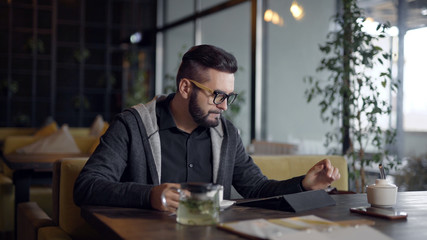 alone young man is resting alone in cafe in daytime, using tablet and sipping hot drink tea from cup