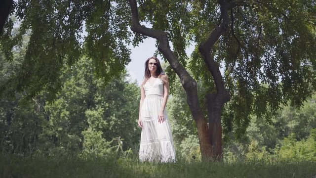 Portrait adorable young girl with long brunette hair wearing sunglasses and a long white summer fashion dress standing under the branches of a tree in the park. Leisure a pretty woman outdoors.