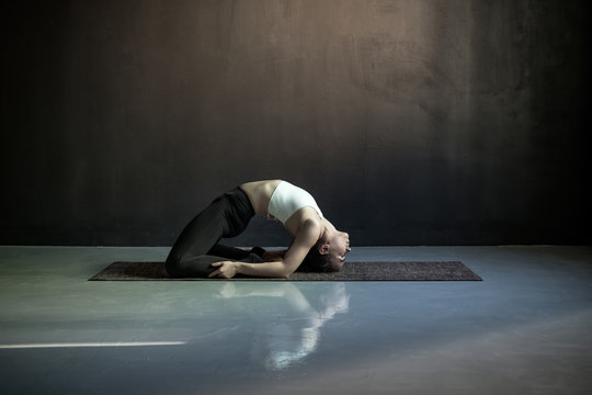 Young woman practicing yoga, doing Pigeon exercise or Kapotasana pose. Indoor full length on black studio wall