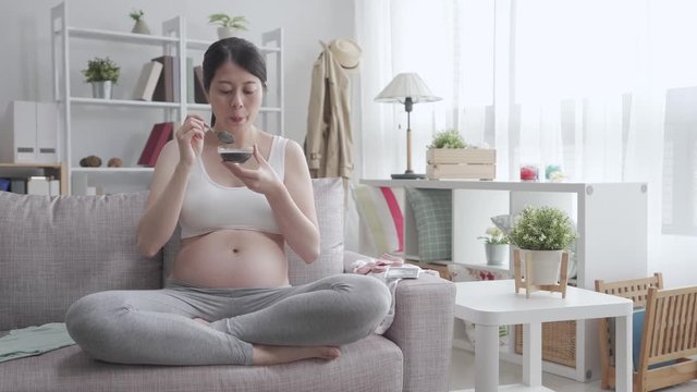 young chinese pregnant lady eating health preserving medicinal cuisine. beautiful mother expecting her baby coming and touching her bare belly while sitting on sofa. future mom enjoying fresh food.