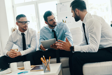 Discussing new project. Group of young modern men in formalwear working using digital tablet while sitting in the office