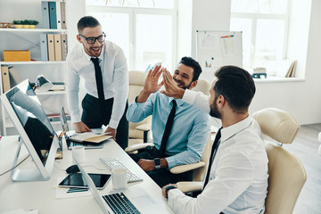 High five for success. Group of young modern men in formalwear smiling while working in the office