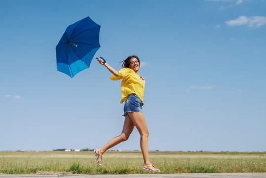 Cheerful Caucasian teenage girl in yellow blouse, denim shorts and with eyeglasses running outdoor with blue umbrella in hand.