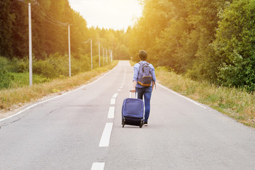 Middle-aged Woman tourist with a travel bag on wheels and a backpack walks along the narrow rural road into the distance. Travel concept