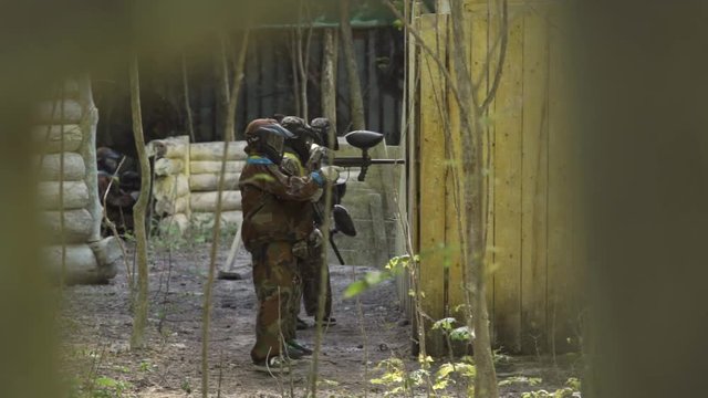 Children and teenagers play paintball in the woods. Several players shoot from behind the building.