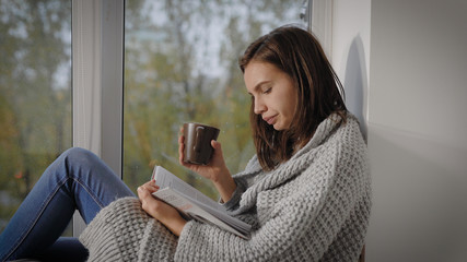 Portrait of a charming brunette girl reading a book at the window with a cup of hot tea, relaxing on weekends.