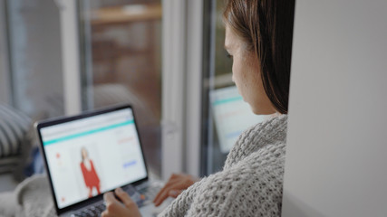 young woman is shopping online, watching pictures, choosing garment on a site using laptop, resting in her flat