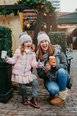 Mother with child girl drinking hot chocolate coffee at Christmas market celebrating New Year holiday. Family outdoor winter activity. Mom and daughter spend time together. Authentic lifestyle.