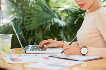 Close-up of young businesswoman writing new ideas with pen in her notepad and using laptop computer in work while sitting at the table in cafe