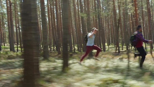 Two frightened young men with backpacks running fast from beast among trees in forest and screaming, tracking right shot