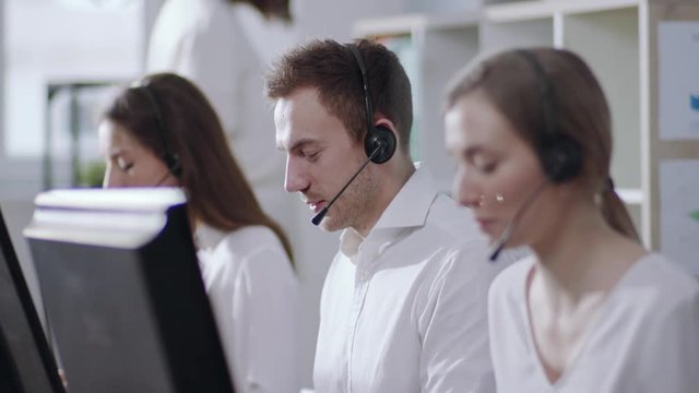 Business and technology concept - helpline operator with headphones in call centre working hard, then looks straight to camera and smiles happily. Politeness, good mood, positive emotions. Female