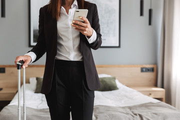 Cropped image of a beautiful young business woman in formal wear clothes indoors at home with suitcase using mobile phone.