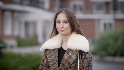 calm pretty brunette is standing alone in cold fall day in yard of houses in city and looking at camera