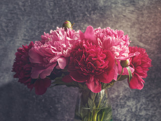 A bouquet of red and pink peonies is in a glass transparent vase. Dark stone background.