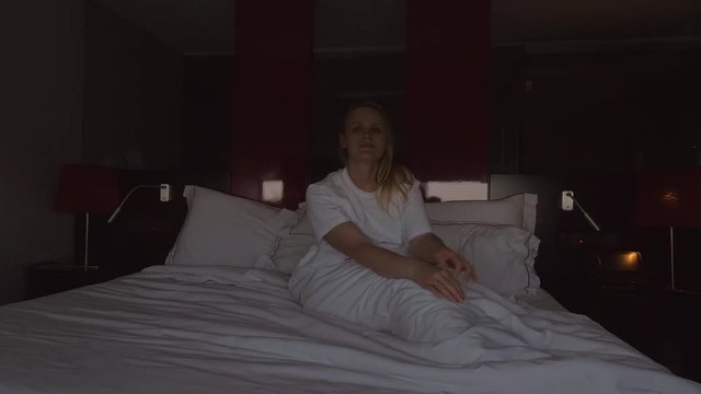 A young woman sits up in a hotel bed, while the blinds are automatically lifted and the bedroom is filling with light
