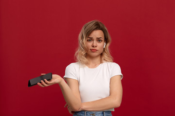 Portrait of a girl with curly blond hair in a white t-shirt on a red background. Unhappy model in wireless headset holds mobile phone.