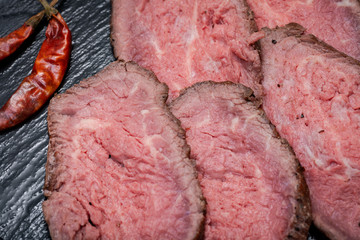 Sliced Grass Fed Juicy Corn Roast Beef garnished with dried Red Chile De Arbol Pepper on black natural stone background.