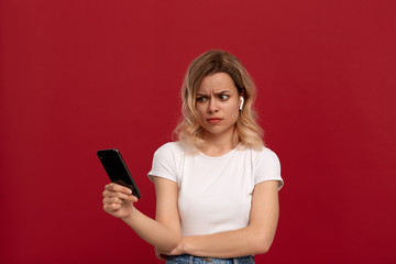 Naklejka premium Portrait of a girl with curly blond hair in a white t-shirt on a red background. Model with dissatisfied look in wireless headset looks at the screen of a mobile phone.