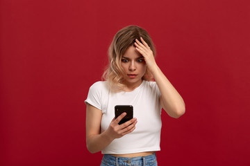 Naklejka premium Portrait of a girl with curly blond hair in a white t-shirt on a red background. Model with dissatisfied look in wireless headset looks at the screen of a mobile phone.