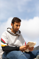 Young Bearded Man Reading A Book Sitting In A Park During The Morning.