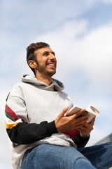 Young Bearded Man Reading A Book Sitting In A Park During The Morning.