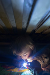 Little child, hiding under the bed, hugging teddy bear and holding flashlight