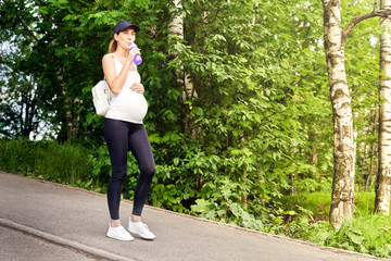A pregnant young woman in a tracksuit and a baseball cap walks in the Park among the greenery and...