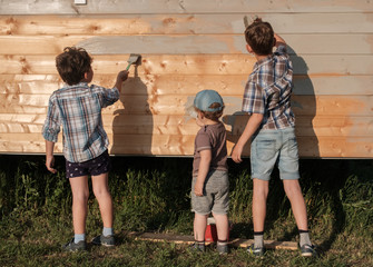 three little boys paint the wall together paint the wall of a wooden house outside. three brothers help to paint new house