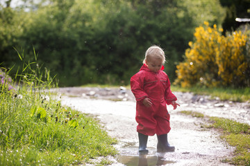 Sweet little toddler boy, blond child in red raincoat and blue boots, playing in the rain in muddy puddles, jumping and laughing