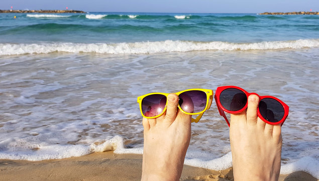 Bare feet raised up with colored sunglasses on the beach. Beautiful sea surf and blue sunny sky in background. Summer vacation. Funny imagination happy character 