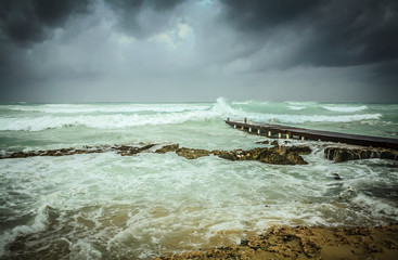 stormy sea waves crashing into wooden pier and big dark grey storm clouds approaching