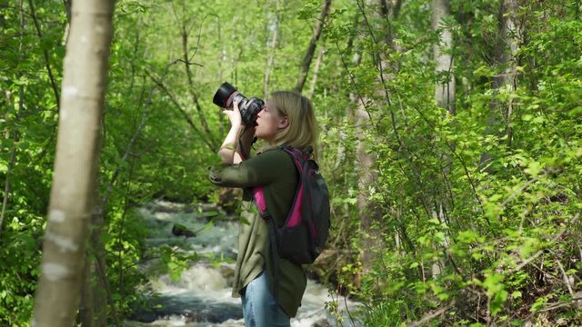 Tracking right shot of female hiker with backpack standing near waterfall in forest and photographing nature with camera