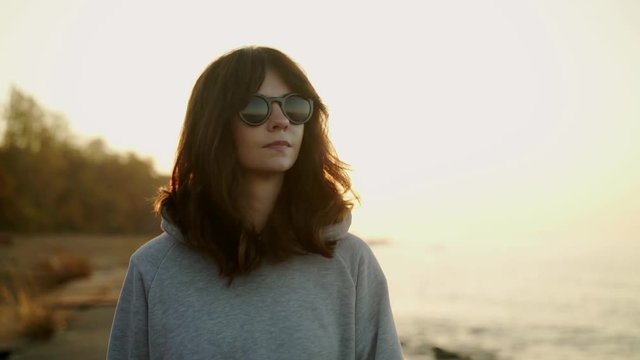 Slow motion of caucasian young woman wearing grey hoodie and sunglasses walking along the beach on background of ocean waves and sky horizon at sunset