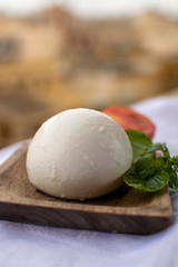 Fototapeta na wymiar Italian mozzarella soft cheese ball served with basil and tomato on wooden plank with yellow houses in old Italian town on background
