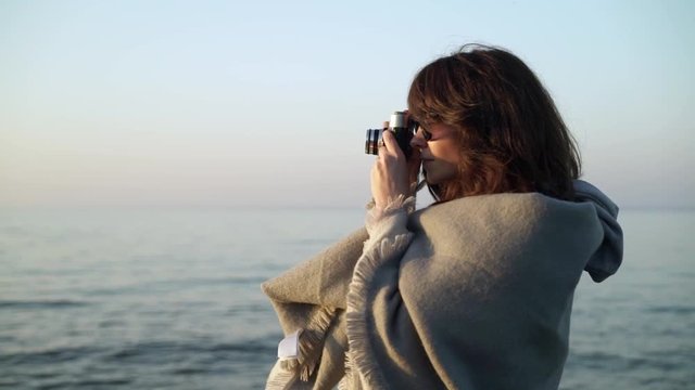 Slow motion of caucasian young woman wearing grey scarf and sunglasses taking photos with film camera of ocean on background of ocean waves 
