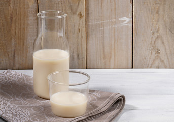 Oat milk in glass bottle and glass on linen napkin on white table on wooden background. 