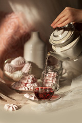 Fototapeta premium A young woman's hand pours boiling water from a teapot into a transparent Cup with fruit tea on a background of pink marshmallow and meringue. Tea party. Tea made from hibiscus flowers.