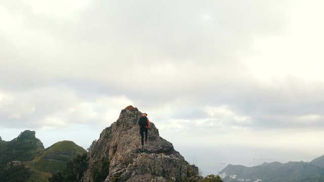 Young woman with backpack climbs a sharp ridge and reaches the top of a mountain. Lady on the top of a mountain among tropical plants in beautiful scenery on Canary Islands
