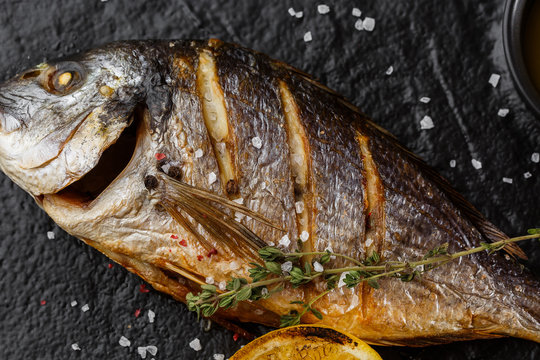 Delicious grilled dorado or sea bream fish with lemon slices, spices, rosemary on dark stone. Grilled sea fish with olive oil, spices and lemon ready for eating. Dorado, herbs and spices. Menu photo
