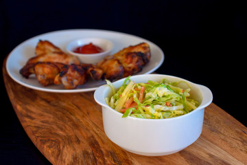 Homemade coleslaw salad in a white plate with chicken wings on a wooden board on a black background
