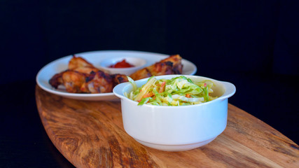 Homemade coleslaw salad in a white plate with chicken wings on a wooden board on a black background