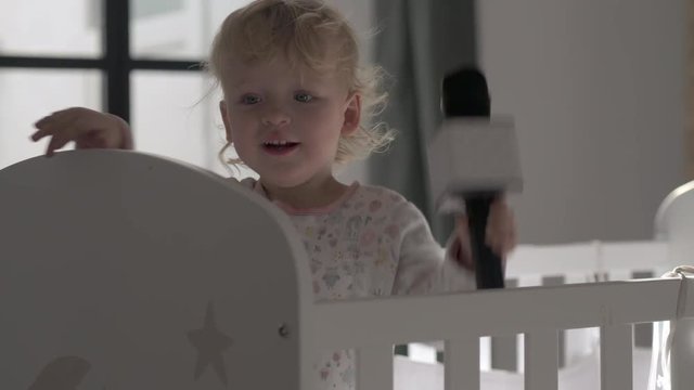 A baby girl, standing in her cot and playing with a toy microphone - sucking and biting it