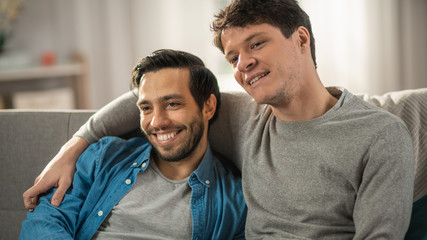 Fototapeta premium Portrait of a Cute Male Queer Couple at Home. They Sit on a Sofa. Partner Embraces His Lover from Behind. They are Happy and Smiling. Room Has Modern Interior.