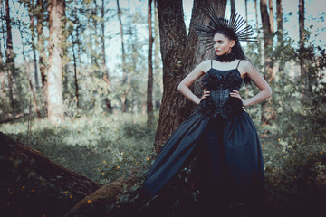 Fototapeta premium elegant woman in witch costume standing on forest background with hands on hips, looking away